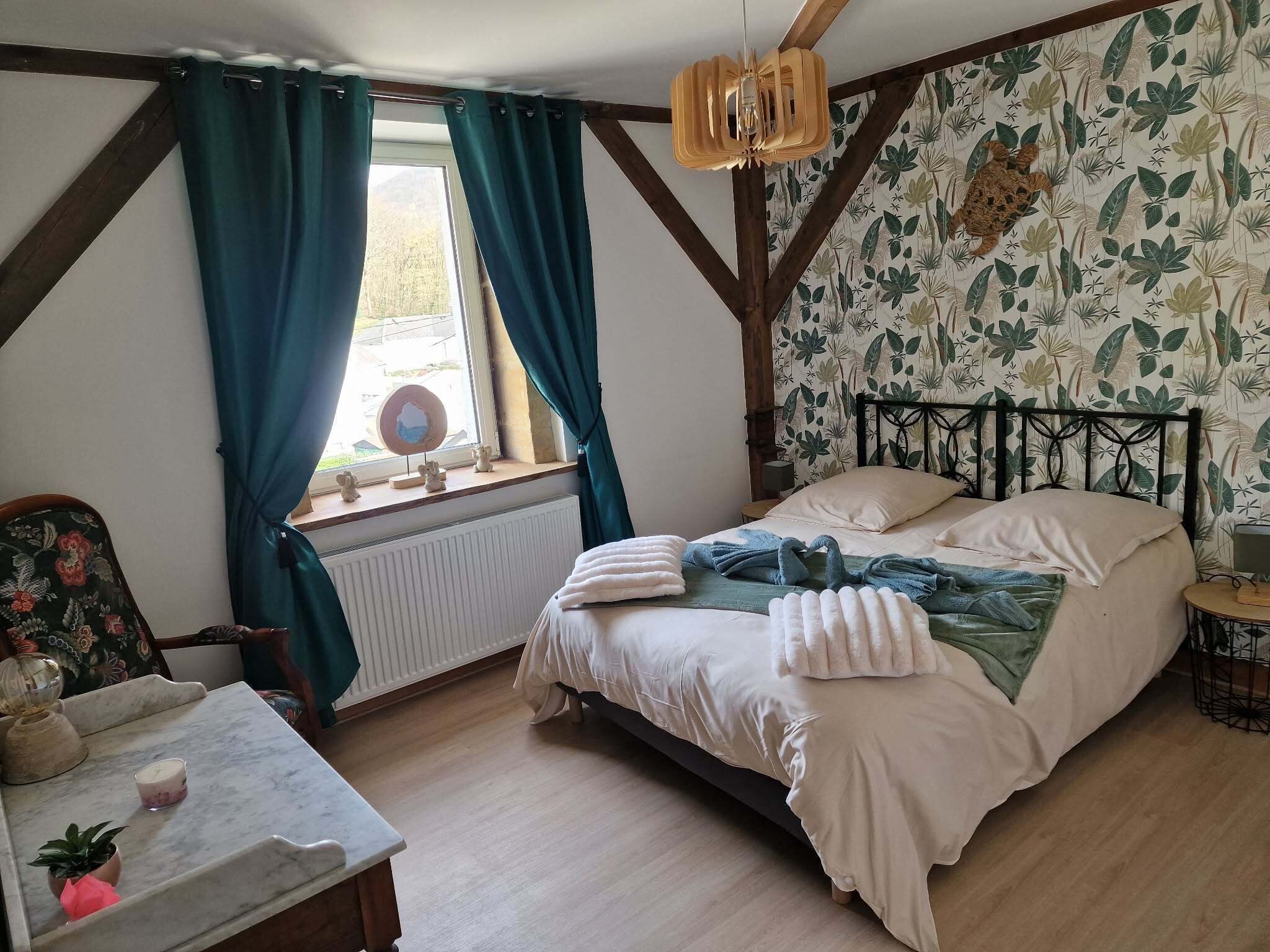 A bedroom with a bed, a window with curtains, a radiator, a small table with a plant, and a wall with a leaf pattern.