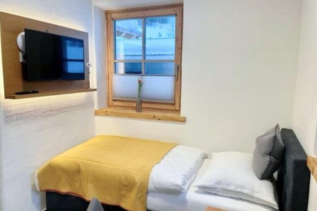 Een kleine kamer met een bed, een televisie bevestigd aan een houten plank, en een raam met jaloezieën.