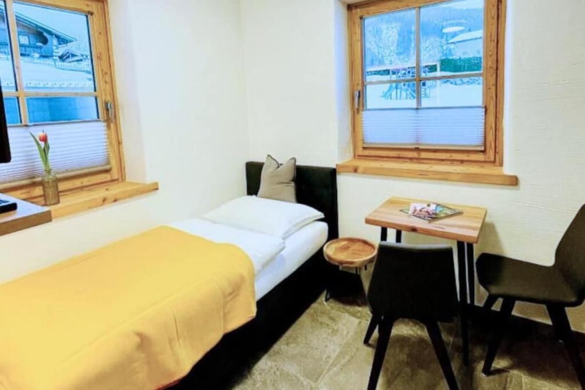 A small room with a bed, a table, two chairs, and a window with a view of a snowy landscape.
