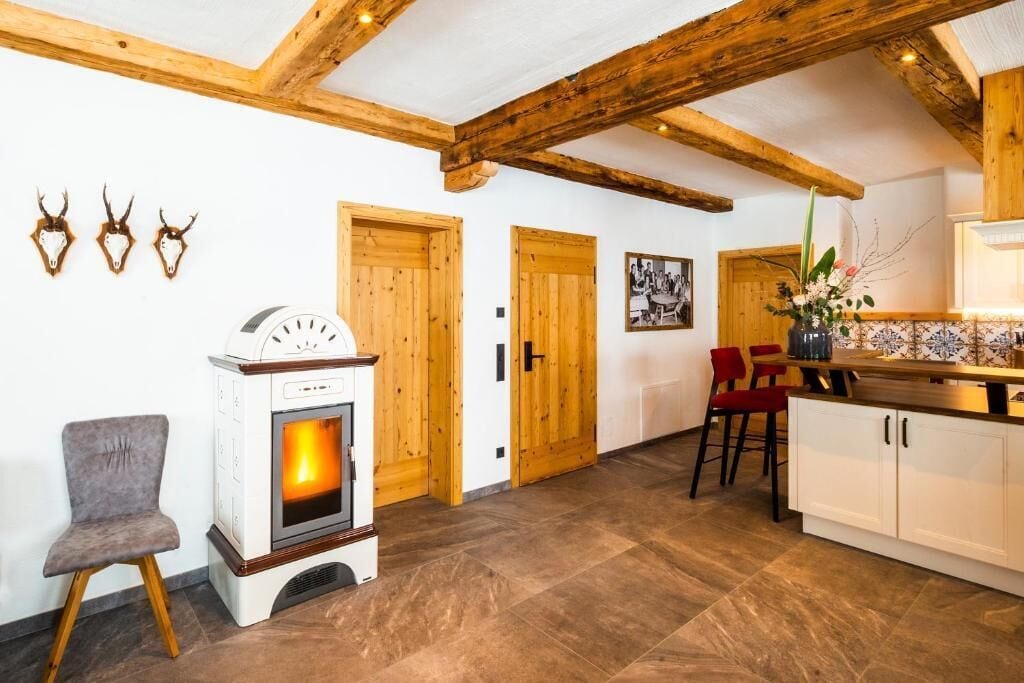 Une pièce dotée d’un poêle à bois, de poutres apparentes, d’un coin repas avec des chaises rouges et d’une cuisine avec des meubles blancs.