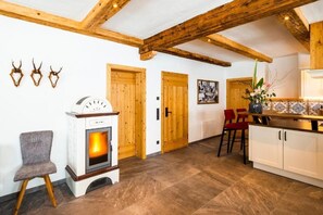 A room with a wood-burning stove, wooden beams, a dining area with red chairs, and a kitchen with white cabinets.
