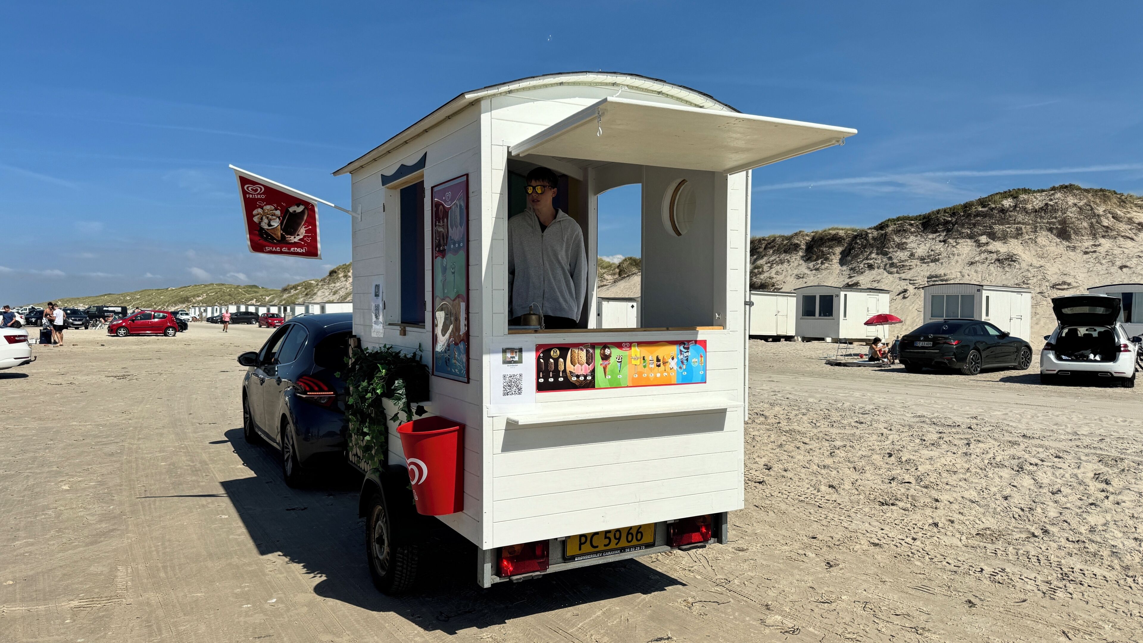 Un stand de vente de nourriture situé sur la plage, avec une façade blanche, un seau rouge et une plaque d’immatriculation.