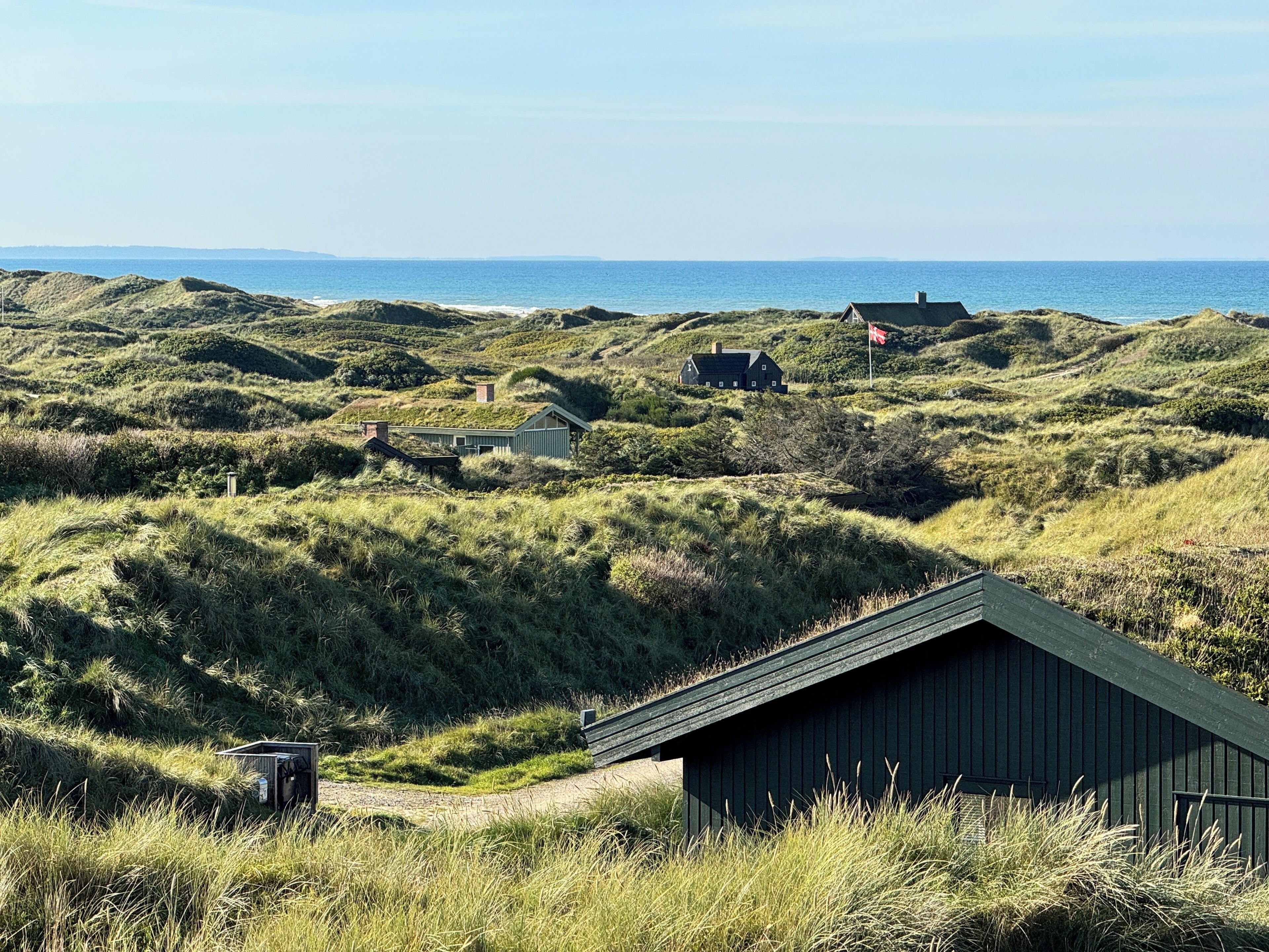 Un paysage côtier avec des dunes herbeuses, une petite construction en bois et une maison surmontée d’un drapeau.