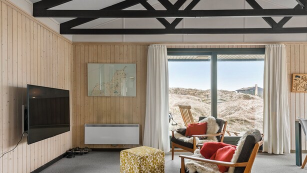 A room with a television, a radiator, a wooden floor, a window with a view of the beach, and a chair with a fur throw. - Cozy 3-bedroom cottage in gorgeous Saltum 7 min. from Denmarks best beach (Saltum)