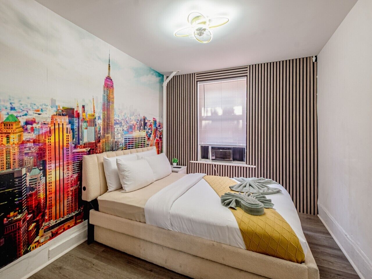 Quarto de hotel com cama, um mural com vista urbana, janela com persianas e ventilador de teto.