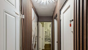 Un pasillo con una puerta blanca a la izquierda, una puerta de madera a la derecha y un techo blanco con una lámpara.