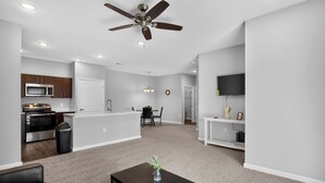 A modern living room with a ceiling fan, a dining area, a kitchen with built-in appliances, and a TV mounted on the wall. - Fresh 1-bedroom layout (Princeton)
