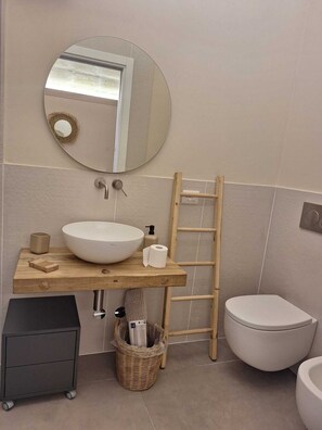 A bathroom with a round mirror, a wooden vanity, a white sink, a toilet, and a small trash bin. - Furnished studio apartment for rent on the ground floor. (Taranto)