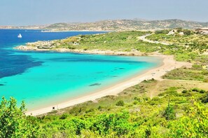 A coastal landscape with a beach, clear turquoise waters, and a hilly backdrop.