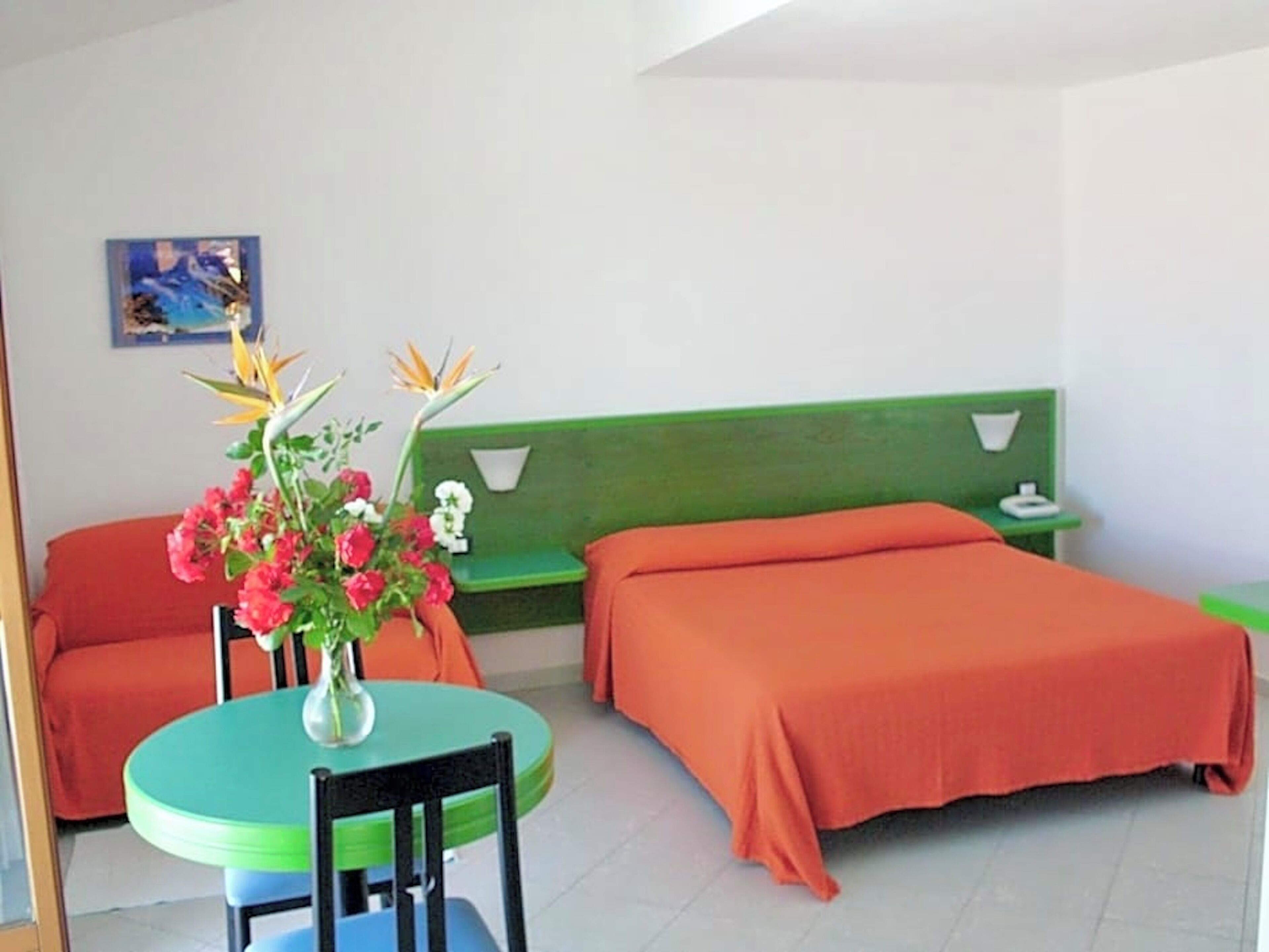 Une pièce avec un lit vert, un lit orange, une table ronde avec une chaise et un vase rempli de fleurs.
