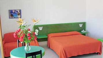 Une pièce avec un lit vert, un lit orange, une table ronde avec une chaise et un vase rempli de fleurs.