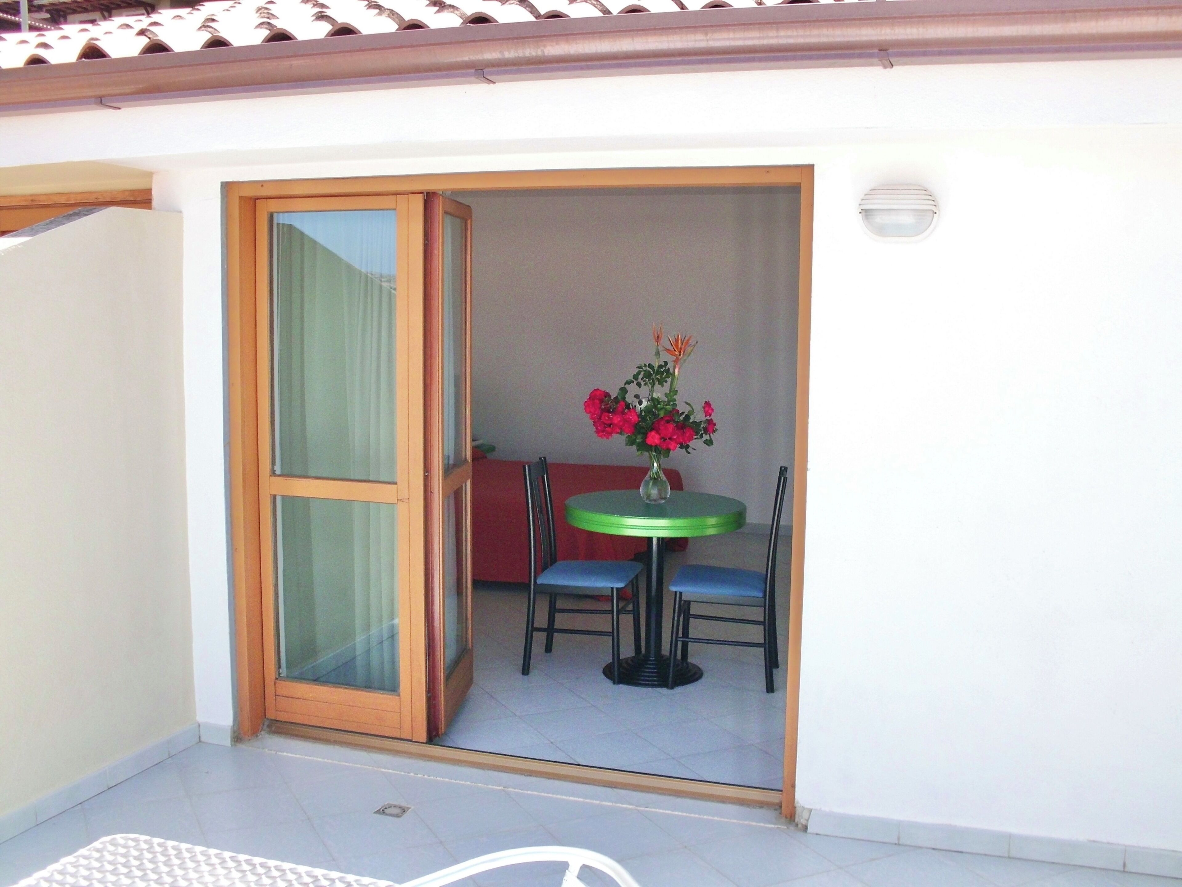 A room with a sliding glass door leading to an outdoor area with a table and chairs, a vase with flowers, and a red sofa.