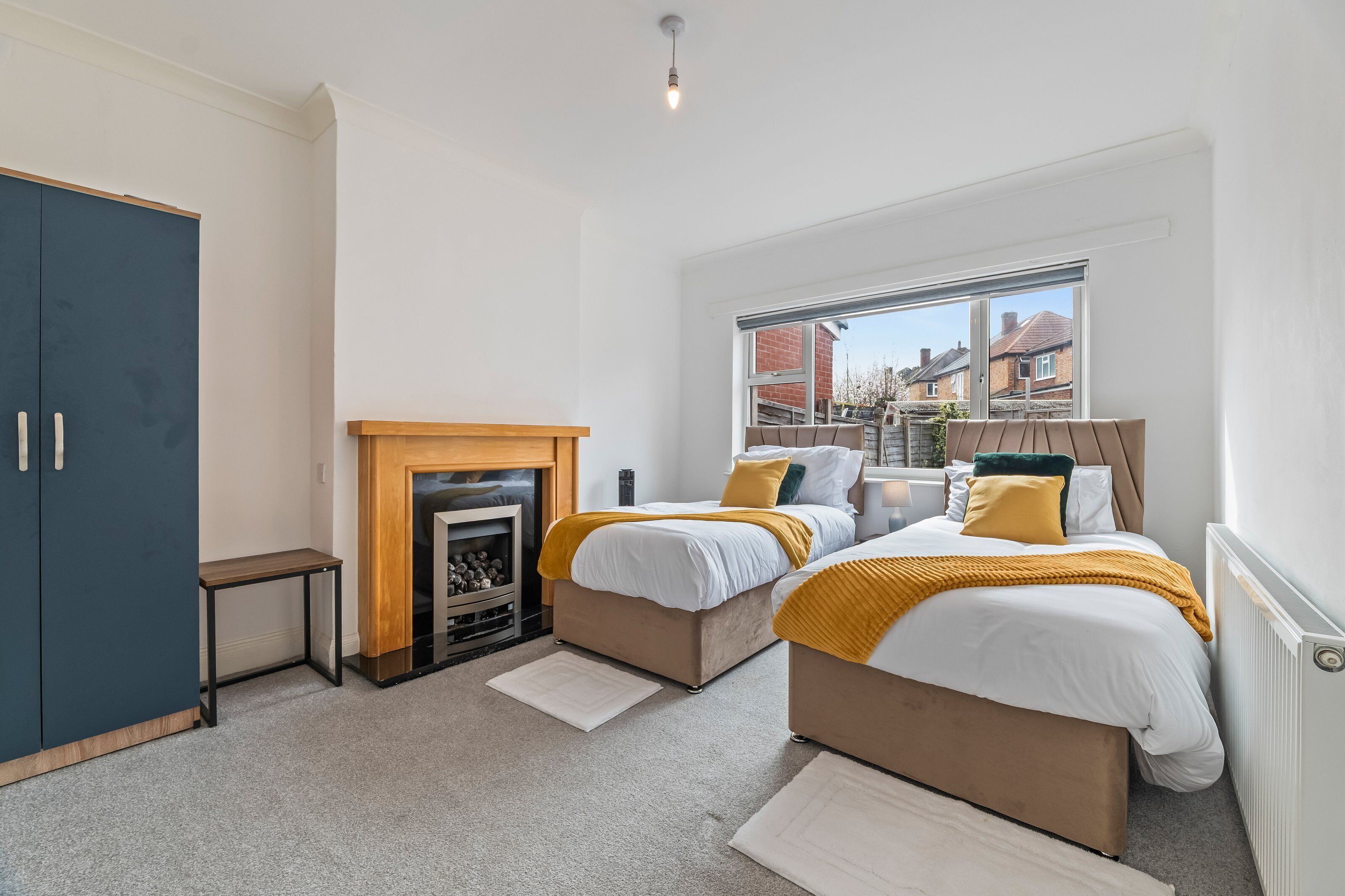A bedroom with two beds, a fireplace, a wardrobe, and a window with a view of houses.
