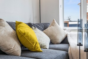 A grey sofa with multiple cushions, including a yellow one, in a room with a window and a view of buildings. - 4-Bed House | Leicester | Driveway Parking (Leicester)