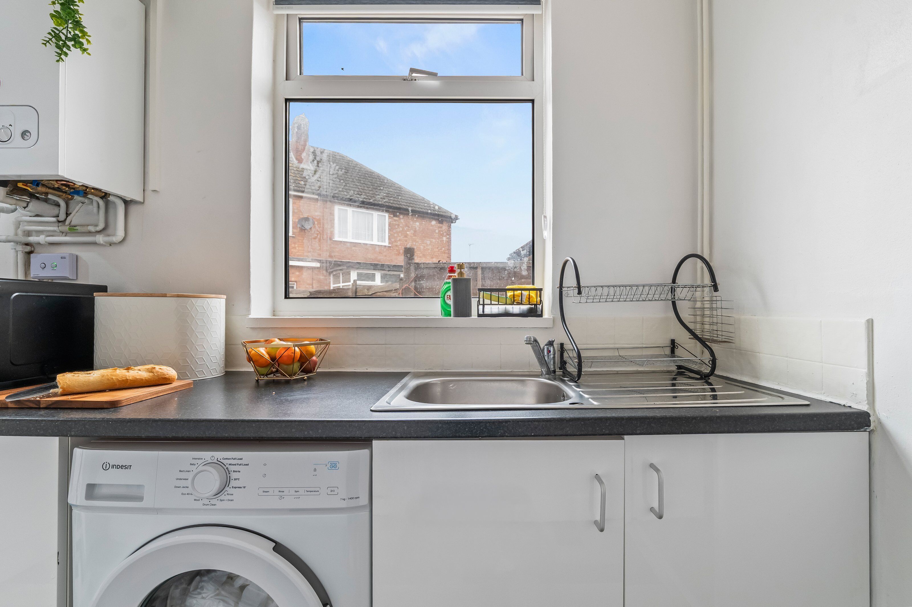 A kitchen with a washing machine, a window with a view of a house, a sink, and a fruit bowl on the counter.