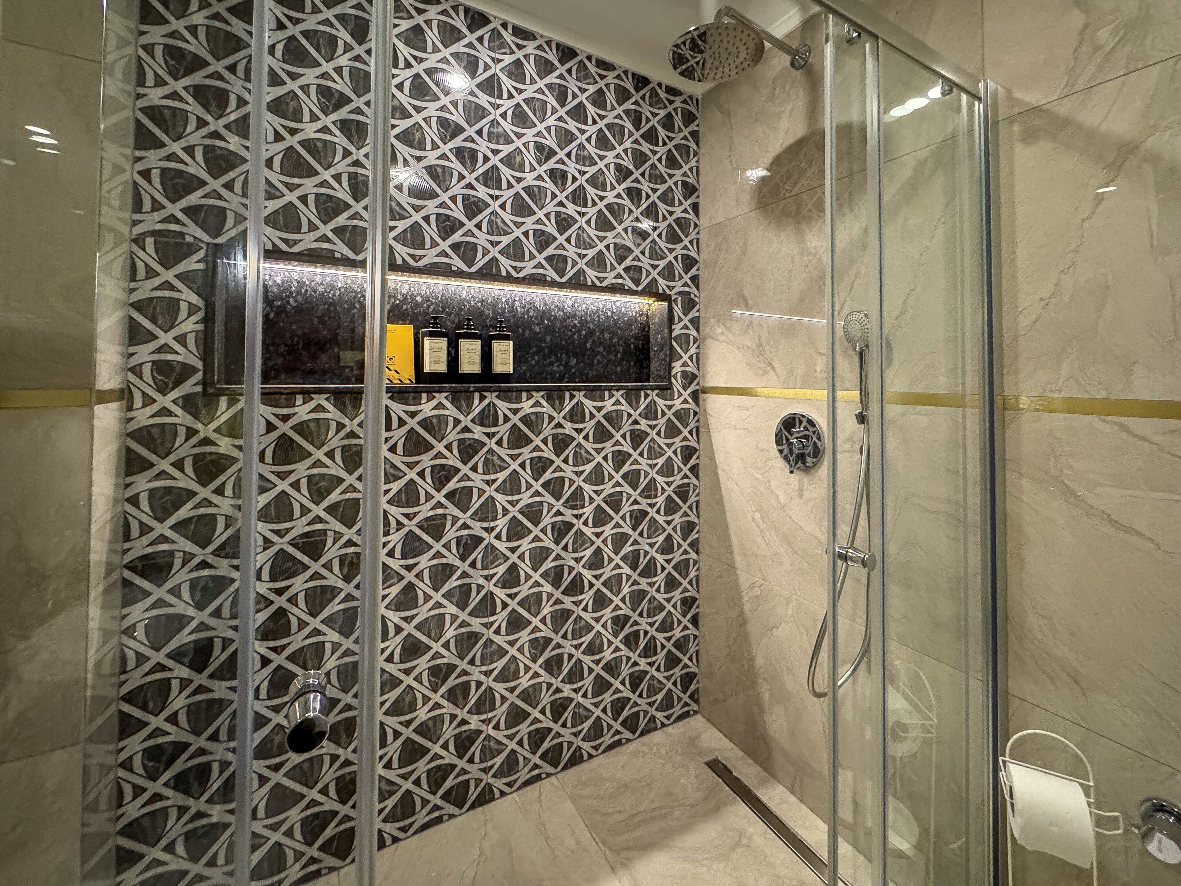 Une salle de bain moderne avec une douche à parois de verre, un mur décoratif orné d’un motif géométrique et une petite étagère contenant des articles de toilette.
