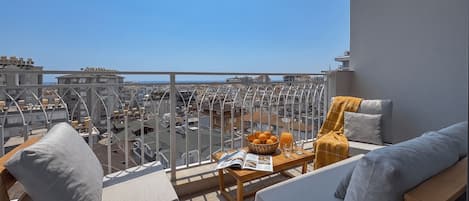Uma varanda com vista para a paisagem urbana e o mar, equipada com um sofá, uma mesa pequena e uma tigela de laranjas.