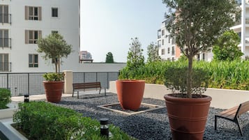 Um jardim no terraço com plantas em vasos, um banco e vista para edifícios vizinhos.