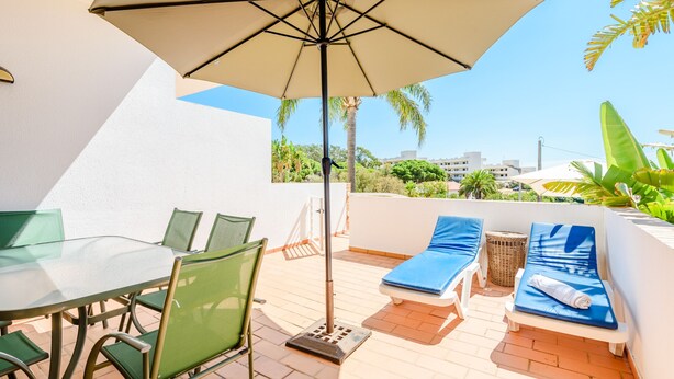 Um pátio com mesa, cadeiras, guarda-sol e espreguiçadeiras.