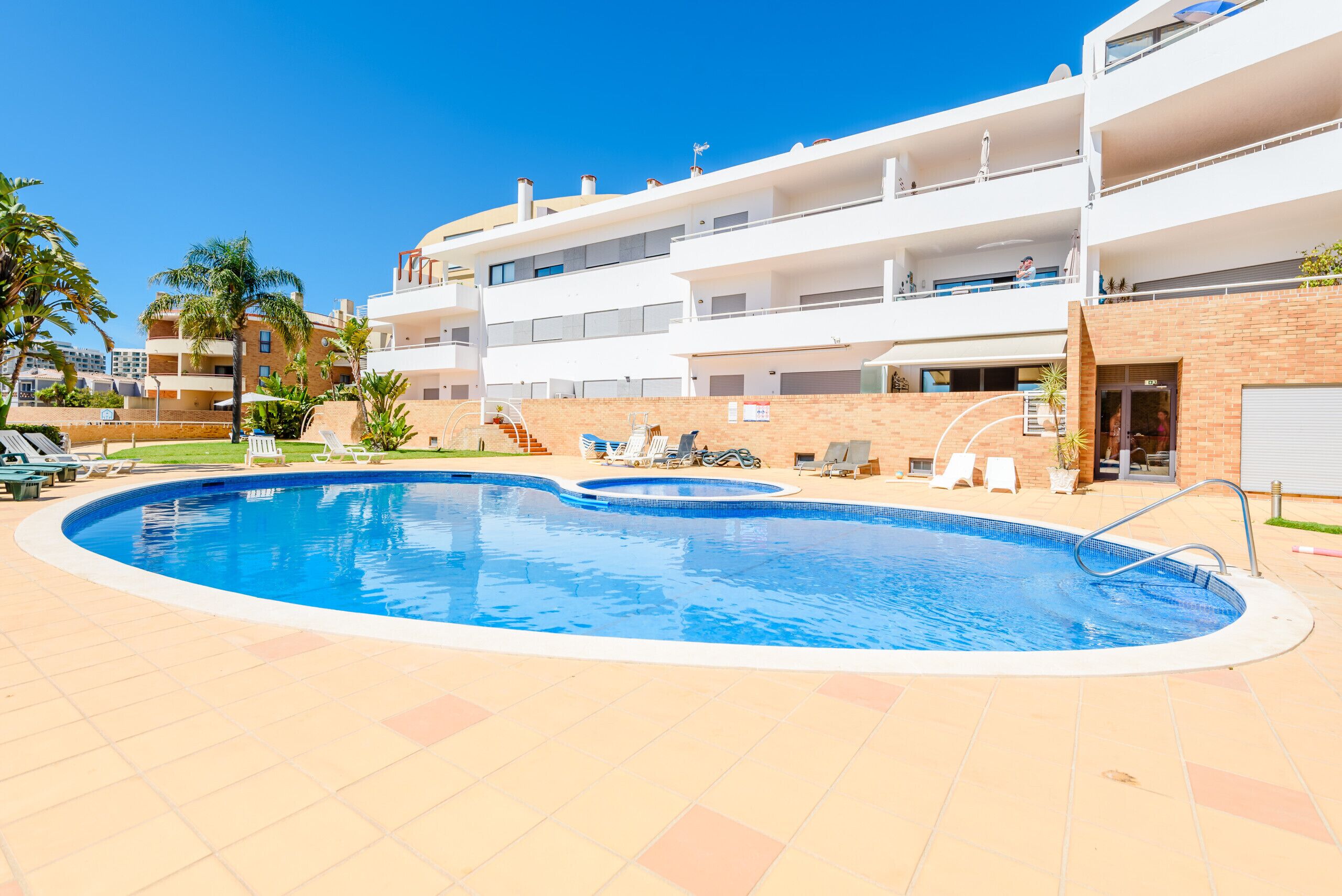 A swimming pool with lounge chairs, a brick wall, and a multi-story building with balconies.
