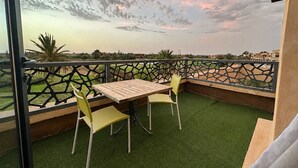 A balcony with a table and two chairs, overlooking a landscaped area with a view of distant buildings. - Exceptional villa overlooking the Amelkis Golf Course - 6 bedrooms, hammam, and indoor pool (Marrakech)