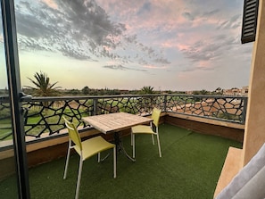 Ein Balkon mit Tisch und zwei Stühlen, der auf einen angelegten Garten mit Blick auf entfernte Gebäude blickt.