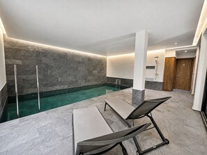 Ein moderner Poolbereich im Innenbereich mit klarem Wasser, zwei Liegestühlen und einer Holztür.