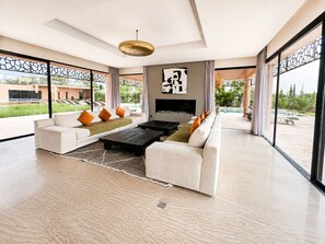 A modern living room with a large sofa, a coffee table, and a fireplace. There is a large window providing a view of the outdoors. - Exceptional villa overlooking the Amelkis Golf Course - 6 bedrooms, hammam, and indoor pool (Marrakech)