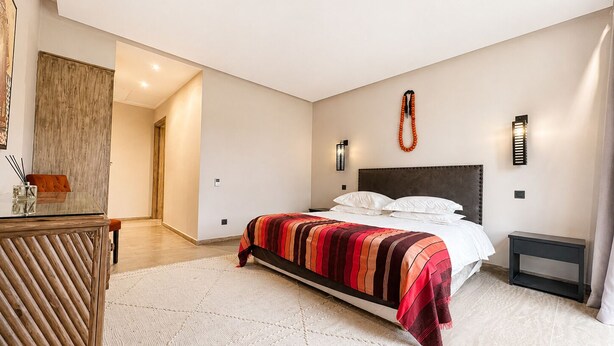 A bedroom with a bed, bedside tables, a nightstand, a wooden wardrobe, and a wall-mounted light fixture. - Exceptional villa overlooking the Amelkis Golf Course - 6 bedrooms, hammam, and indoor pool (Marrakech)