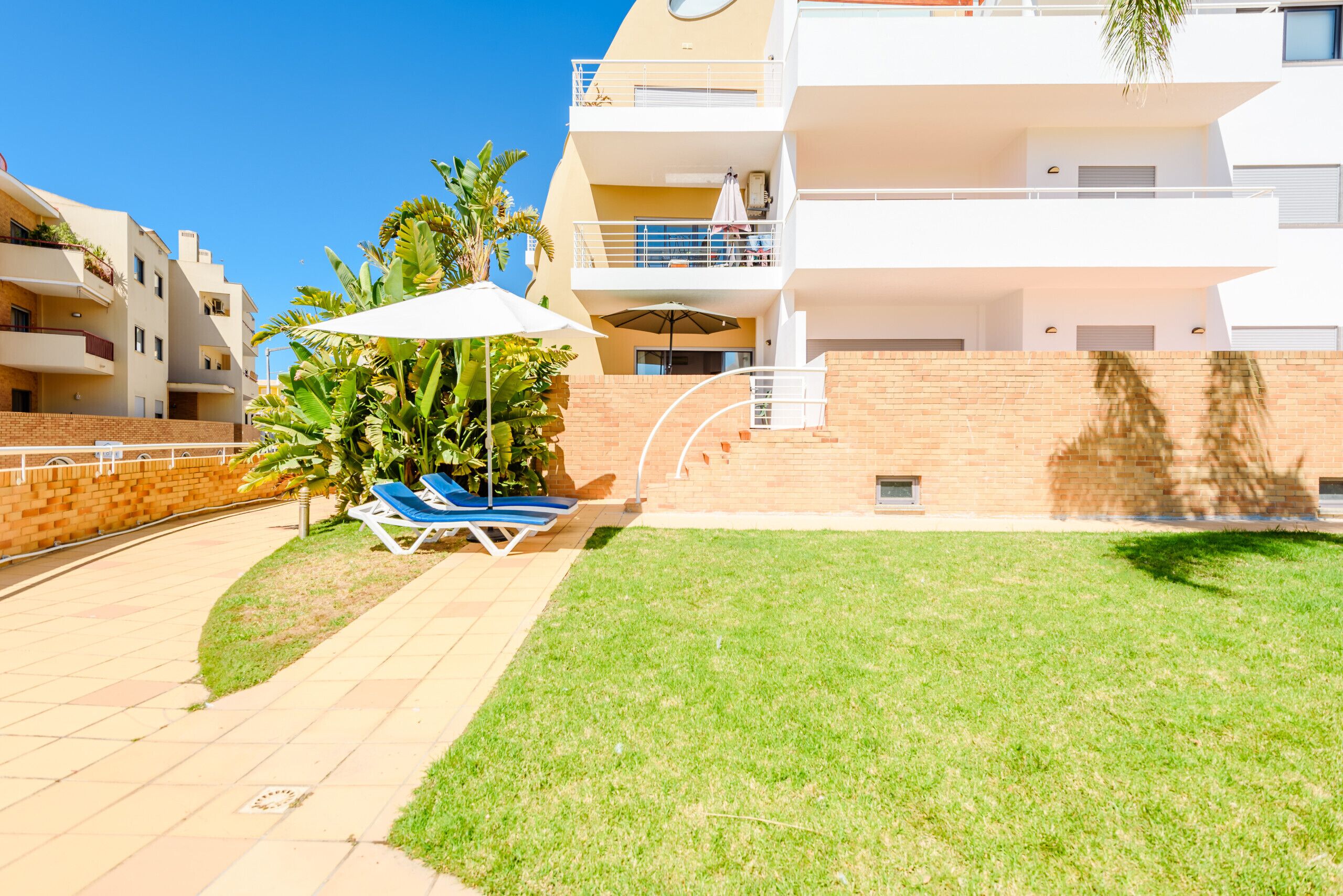 A multi-story residential building with a flat roof, a brick wall, and a small grassy area with a white umbrella and two lounge chairs.