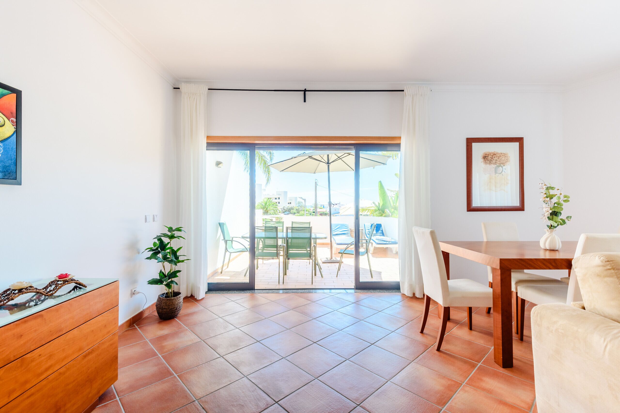 A room with a dining table, chairs, a potted plant, and a view through a sliding glass door to an outdoor area with a table and chairs.