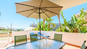A glass-top table with four chairs, a large beige umbrella, and lush tropical plants. - Apartment Praia Dona Ana (Lagos)