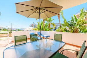 A glass-top table with four chairs, a large beige umbrella, and lush tropical plants. - Apartment Praia Dona Ana (Lagos)