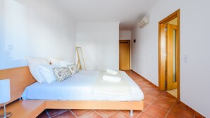 A bedroom with a bed, a bedside table, a lamp, a wooden door, and a tiled floor. - Apartment Praia Dona Ana (Lagos)