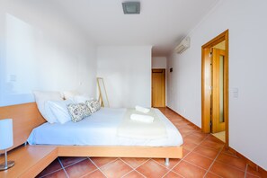 A bedroom with a bed, a bedside table, a lamp, a wooden door, and a tiled floor. - Apartment Praia Dona Ana (Lagos)