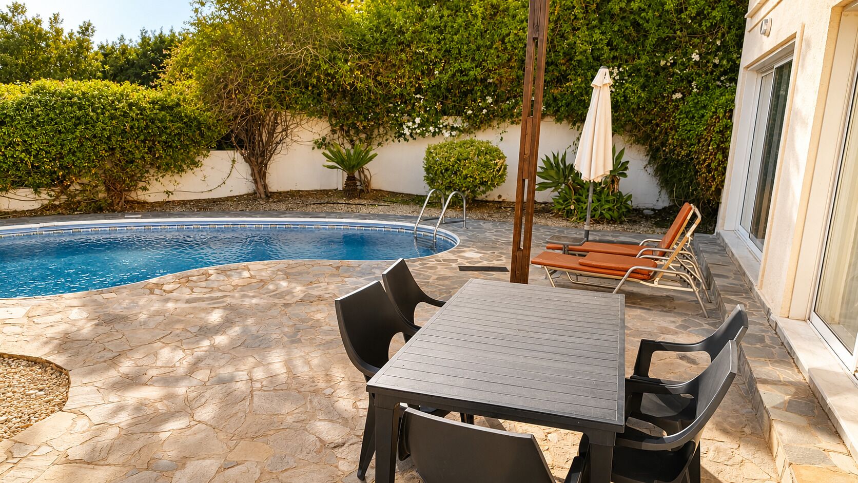 A poolside area with a table and chairs, a wooden umbrella, and a stone patio.