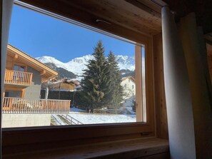 Ein Fenster mit Blick auf einen Berg, ein Holzhaus und verschneite Erde.
