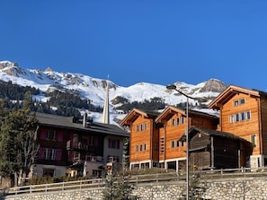 Eine Berghütte mit Holzhütten und einer Steinfassade, vor einer Kulisse schneebedeckter Gipfel.