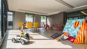 Un moderno vestíbulo de hotel con ventanas amplias, sillas amarillas y rojas, un pequeño coche de juguete y una decoración colorida con una carpa.