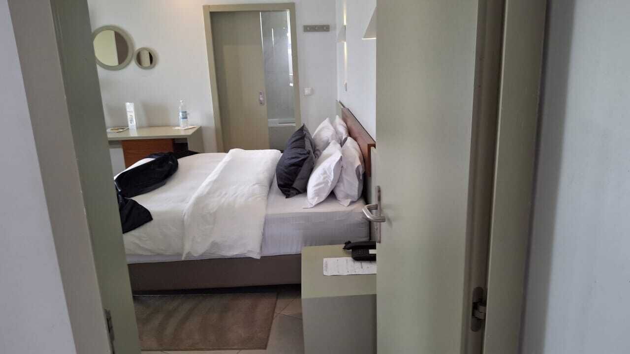 Quarto de hotel com cama, mesa de cabeceira com luminária, espelho e porta que dá para outro cômodo.