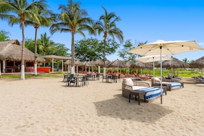 A beachside resort with thatched-roof structures, lounge chairs, and umbrellas. - Coastal Condo with Rooftop Infinity Pool & Beach Club – La Cruz, Nayarit (Bahia de Banderas)