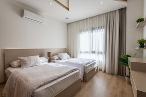 Quarto de hotel com duas camas, mesa de cabeceira, luminária, ar condicionado, janela com cortinas e uma pequena planta em uma prateleira.