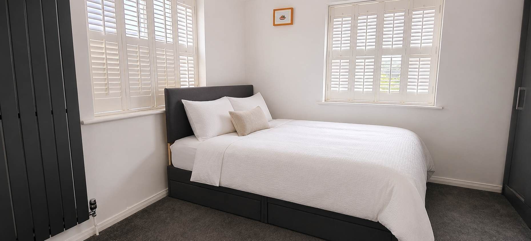 Une chambre à coucher avec un lit, deux fenêtres avec des volets, un radiateur et un tableau accroché au mur.