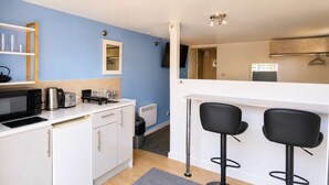 Eine kompakte Küche mit weißen Schränken, einer Mikrowelle, einem Wasserkocher und einem Toaster. Zwei schwarze Barhocker und ein kleines Fenster sind ebenfalls vorhanden.