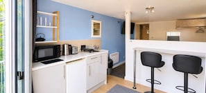 Eine kompakte Küche mit weißen Schränken, einer Mikrowelle, einem Wasserkocher und einem Toaster. Zwei schwarze Barhocker und ein kleines Fenster sind ebenfalls vorhanden.