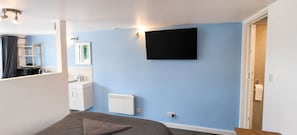 Ein Schlafzimmer mit einem Bett, einem an der Wand montierten Fernseher, einer Heizung und einem durch eine offene Tür einsehbaren Badezimmer.