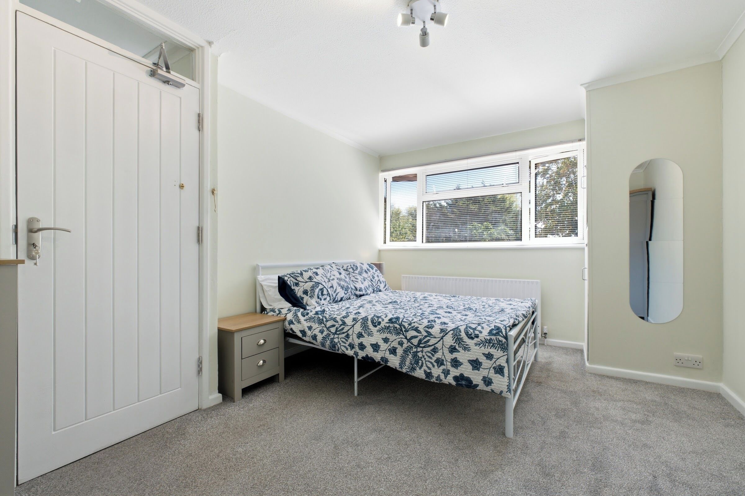 A bedroom with a single bed, a nightstand, a window with blinds, and a door with a handle.