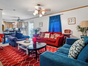 Ein modernes Wohnzimmer mit einem roten Sofa, blauen Sesseln und einem Couchtisch. Im Hintergrund befindet sich eine Küche mit Holzschränken und einem Essbereich.