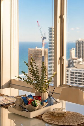 A room with a view of the ocean and a construction crane, a plant on the windowsill, and a table with snacks and magazines. - Luxury Suite| Surfers Paradise|Ocean Views (Surfers Paradise)