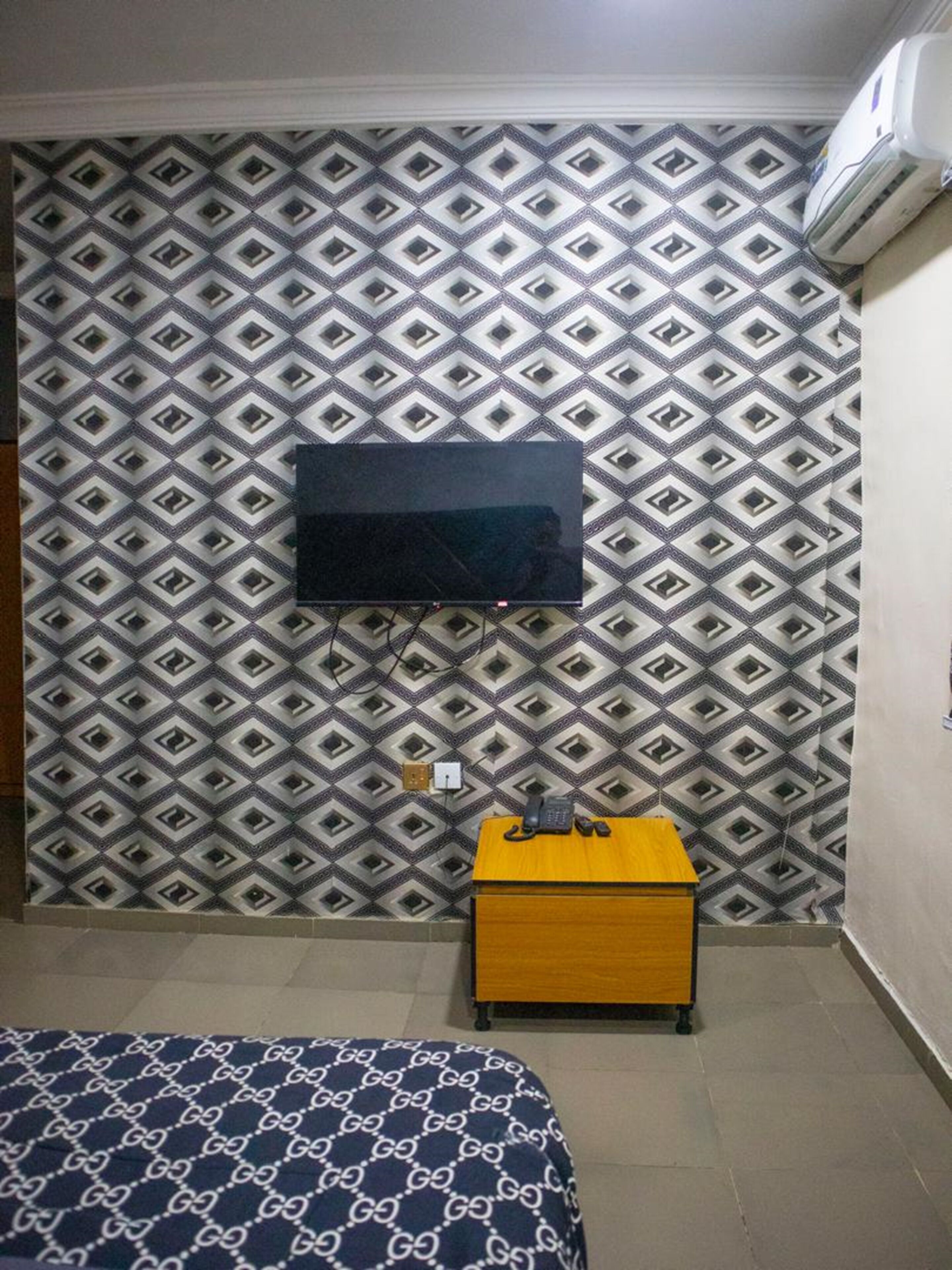 Una habitación con un papel tapiz estampado, un televisor de pantalla plana montado en la pared, una mesita de noche de madera con un teléfono y una cama con colcha estampada.